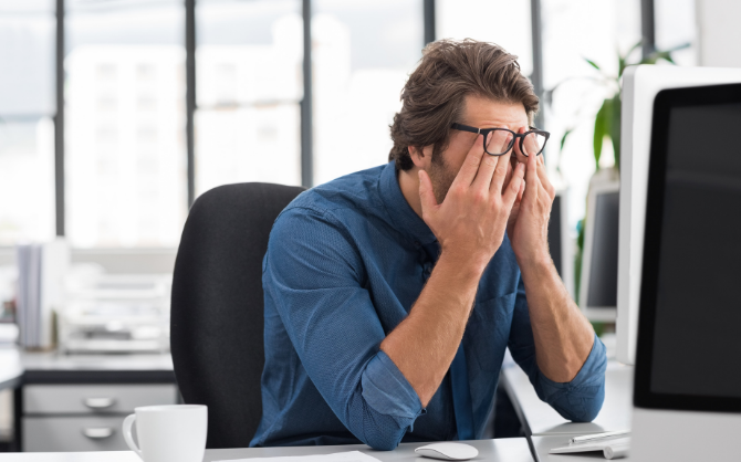 A person in a blue shirt sits at a desk in front of a computer, rubbing their eyes, clearly showing signs of burnout. The bright office space with large windows offers little reprieve. A coffee cup on the table hints at their struggle to stay alert amidst the stress.