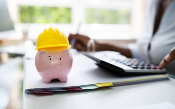 A pink piggy bank wearing a yellow construction helmet sits on a stack of documents with colored tabs