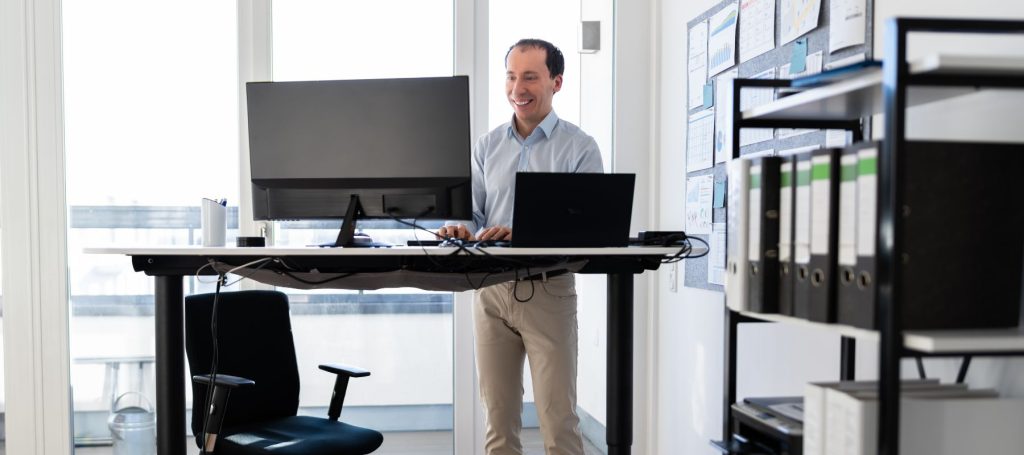 A man stands and works at a height-adjustable desk with monitors and a laptop in a bright, modern office. An empty chair and shelves with binders are visible in the background.