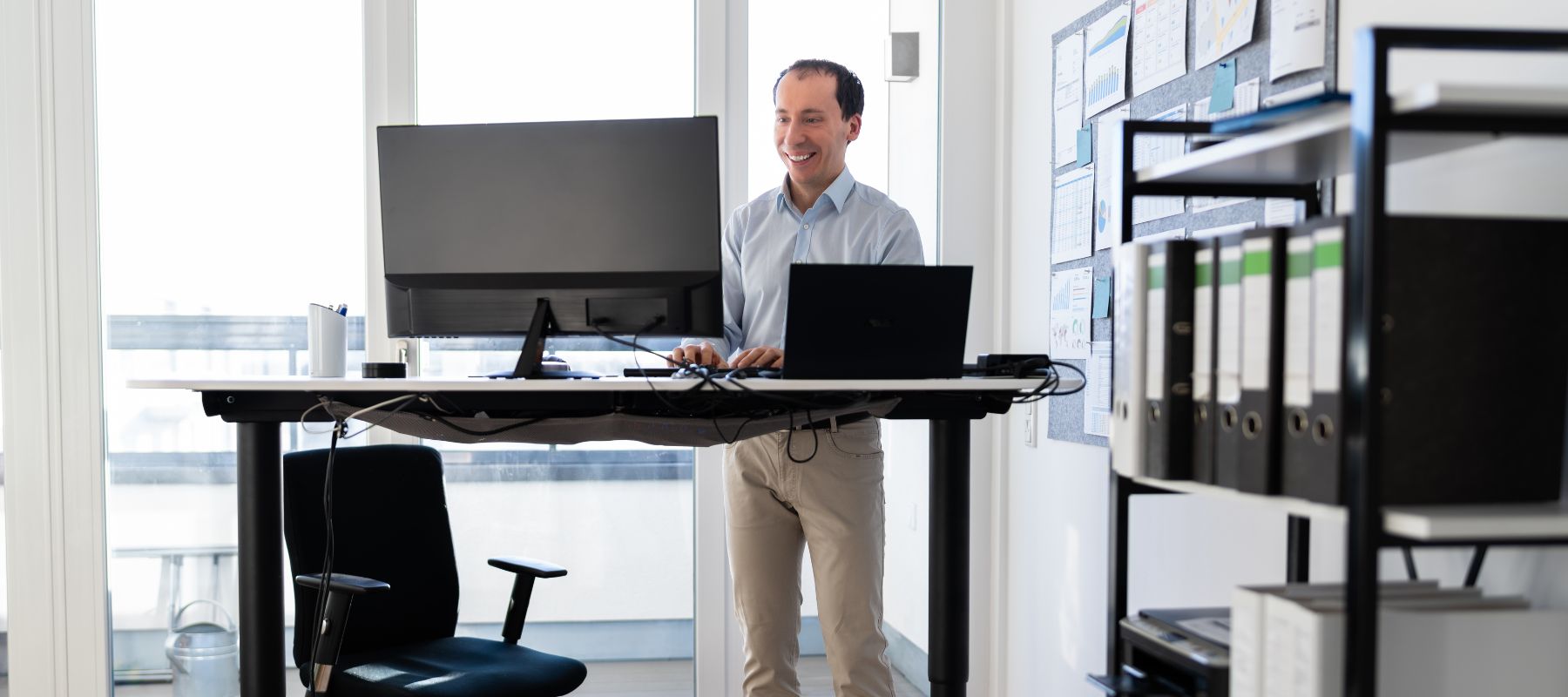 A man stands and works at a height-adjustable desk with monitors and a laptop in a bright, modern office. An empty chair and shelves with binders are visible in the background.
