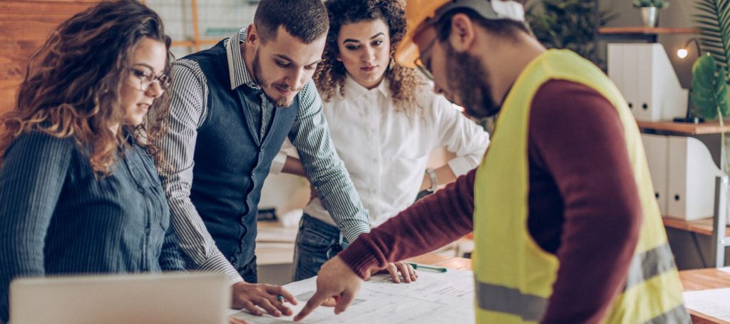 Four people gather around a table reviewing blueprints. One person in a safety vest and hard hat highlights safety risks, while the others, dressed in office attire, observe closely during a WHS consultation in a modern office setting.