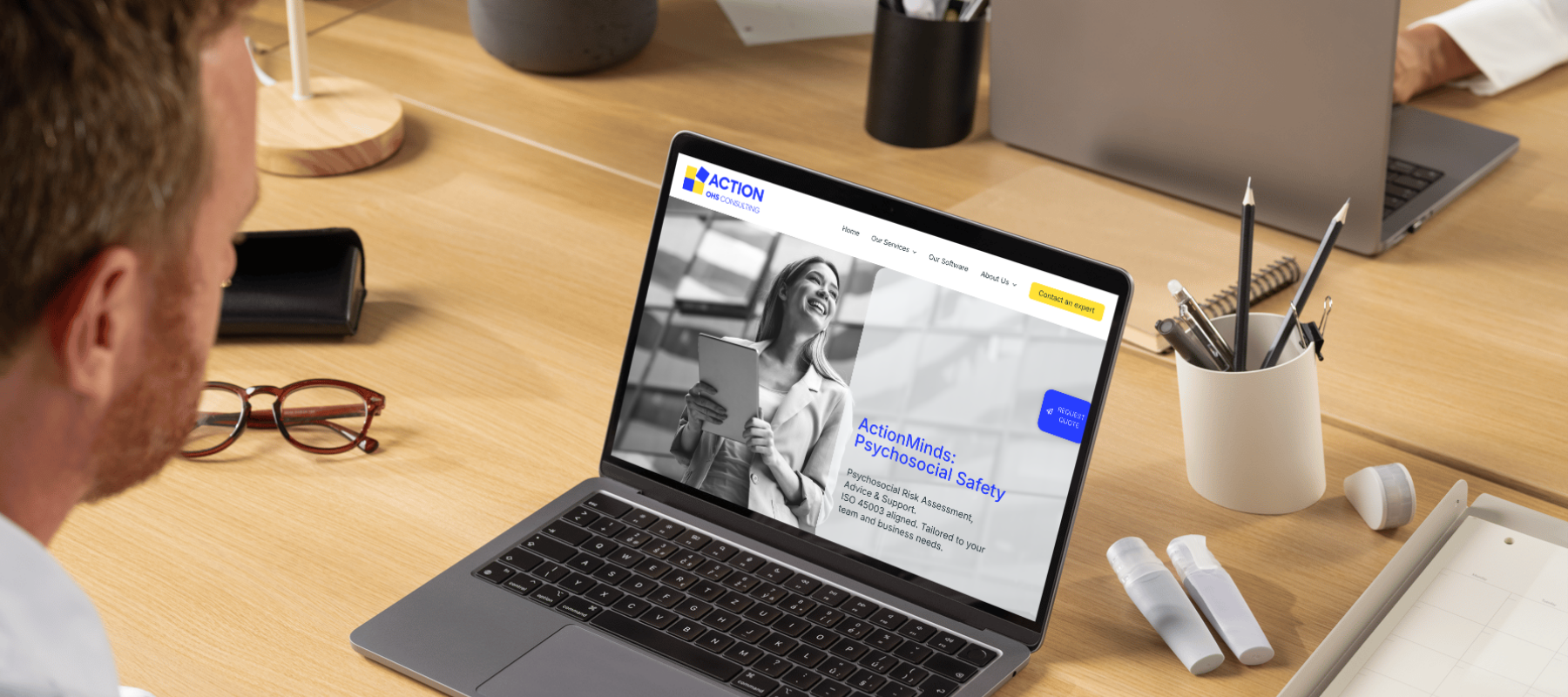A man sits at a wooden desk using a laptop to search for "Psychosocial Safety" with content on psychosocial risk assessment. ActionMinds