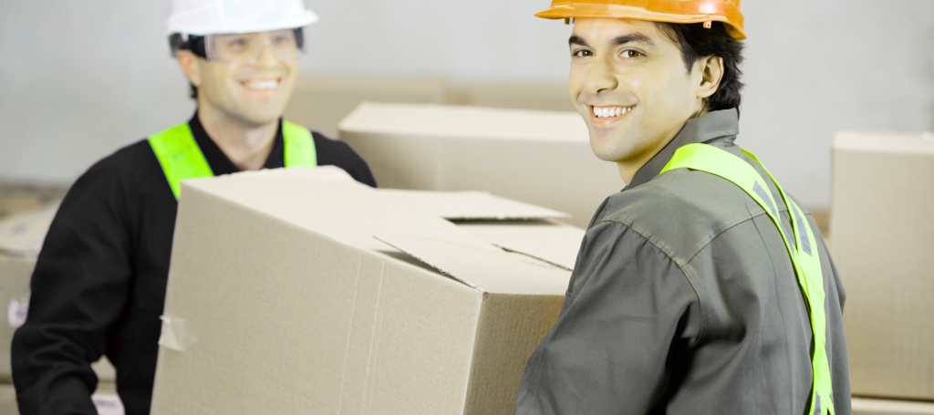 Two workers wearing safety vests and hard hats smile while carrying large cardboard boxes in a warehouse, demonstrating workplace safety and a positive psychosocial environment.