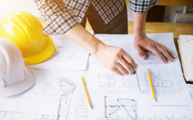 A person in a plaid shirt works on architectural blueprints at a desk, using a pencil and ruler. Two construction helmets, one yellow and one white, underscore the focus on maintaining a safe construction environment.