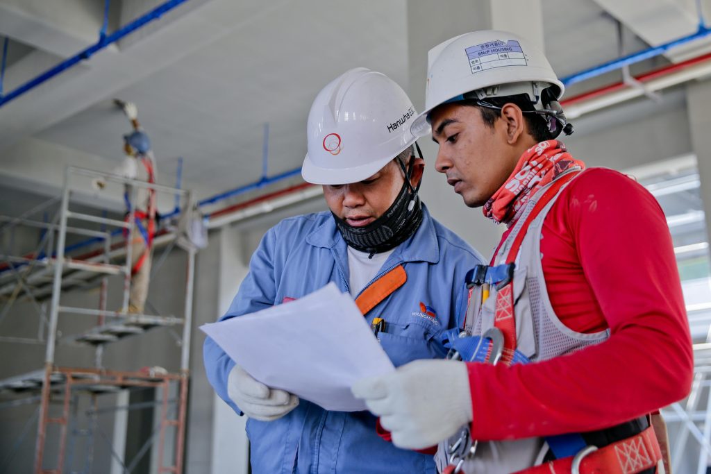 Two construction workers wearing helmets and safety gear review updated plans in an unfinished building. Scaffolding is visible in the background, and one worker is wearing a blue jacket while the other has on a red shirt.