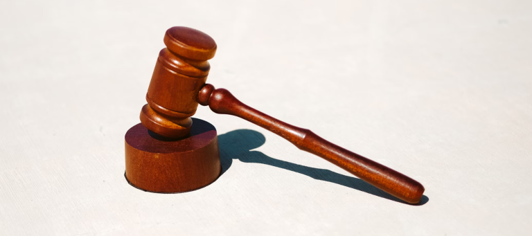 A wooden judge's gavel rests on top of a matching sound block, casting a shadow on a light surface. The gavel is angled slightly, emphasizing the rounded head and handle.