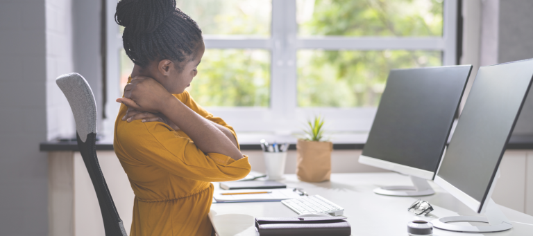 A woman in a yellow blouse practices ergonomics, stretching her neck at a desk with two monitors. Her workspace includes a keyboard, mouse, glasses, and a small potted plant. A large window behind her reveals lush greenery outside.